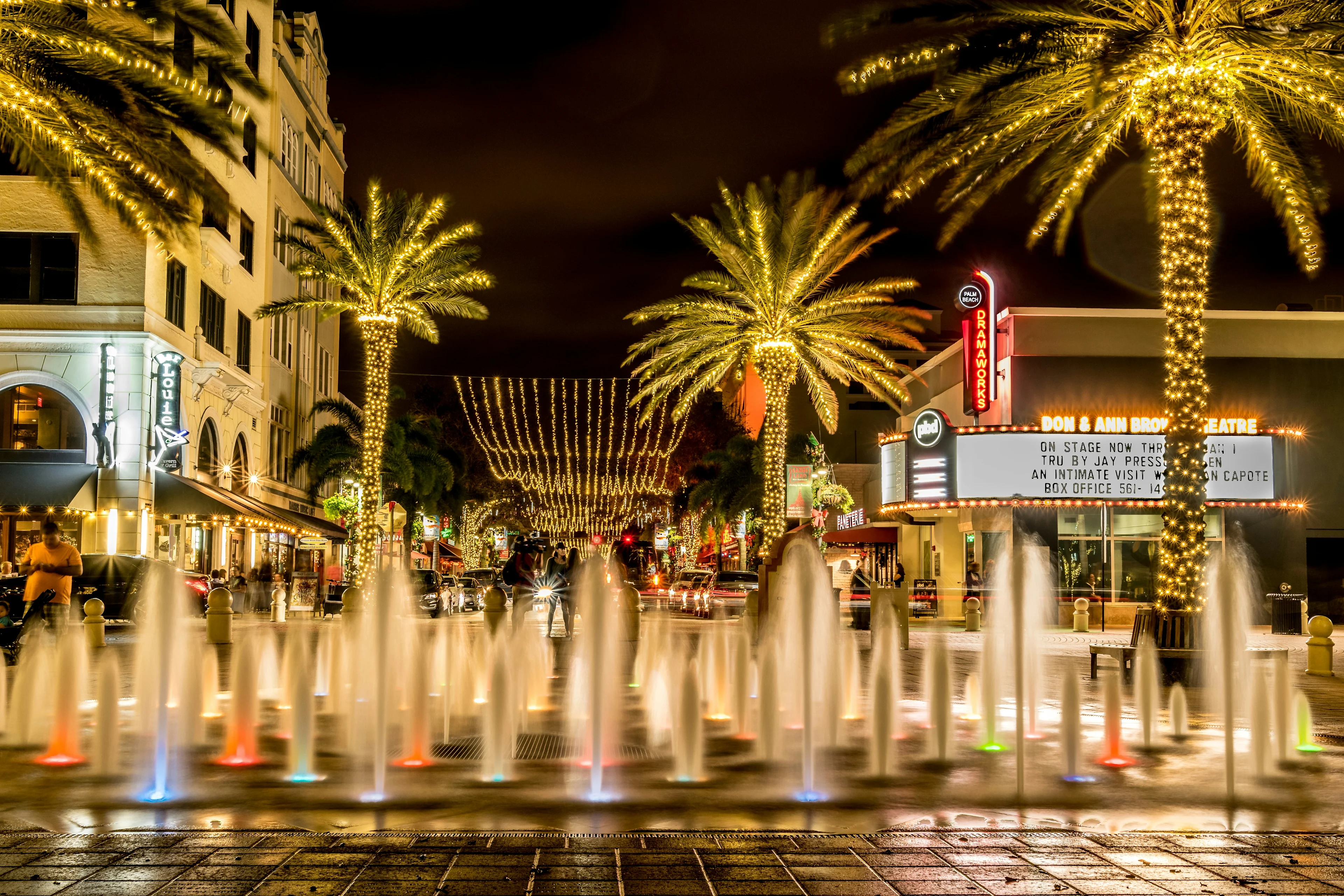 Illuminated fountains and lit palm trees along a downtown West Palm Beach street at night