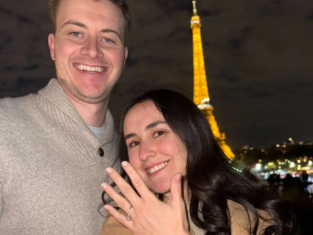 A couple posing at night with the illuminated Eiffel Tower behind them, the woman showing off an engagement ring.