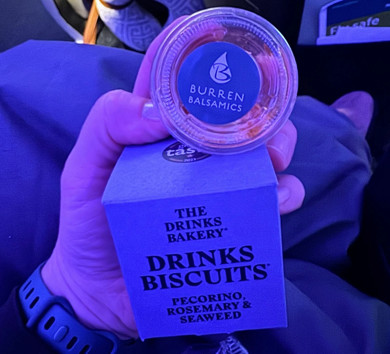 Close-up of a hand holding a Burren Balsamics jar and a box of Drinks Biscuits snack in blue-tinted airplane cabin lighting.