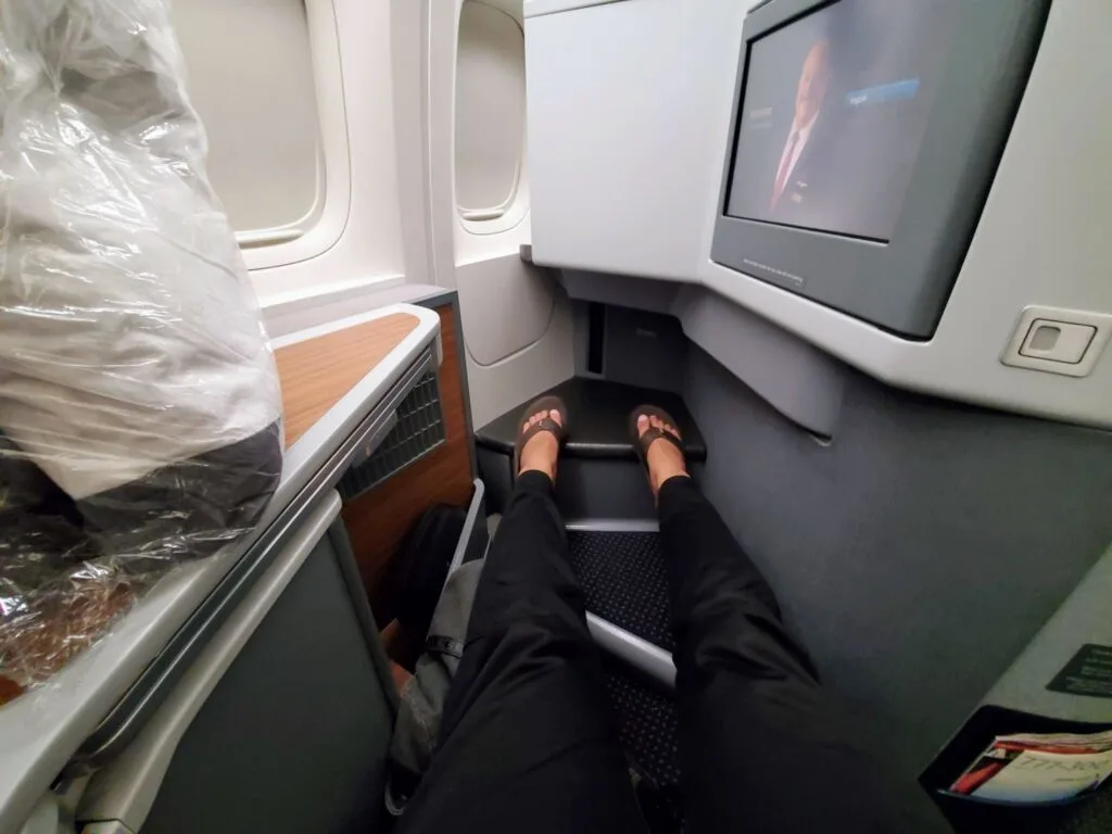 A passenger's legs stretched out in an American Airlines business class seat, with the entertainment screen playing in the background.