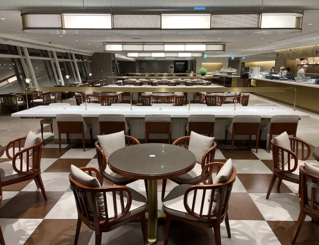 Dining area of the Japan Airlines First Class Lounge at Tokyo Haneda Airport, with wooden armchairs around round tables on a checkered floor
