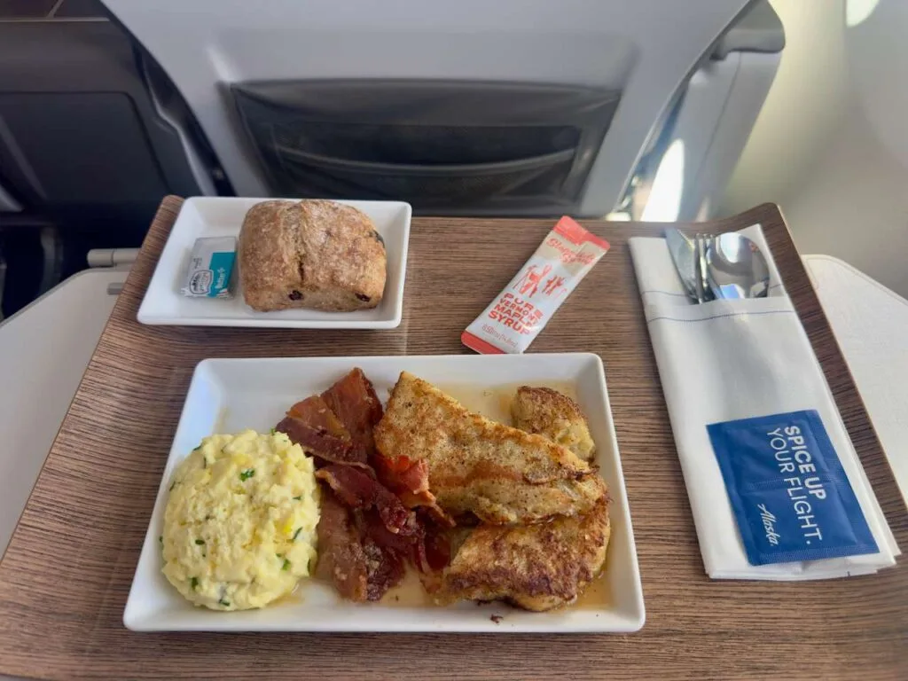 Alaska Airlines first class breakfast tray with scrambled eggs, bacon, French toast, and a bread roll.
