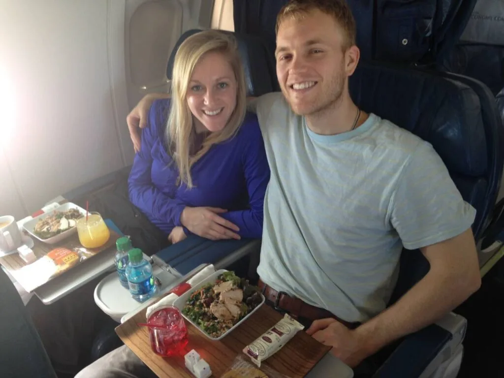 A couple smiling together in airline business class seats with meal trays of salad, fruit, and drinks in front of them.