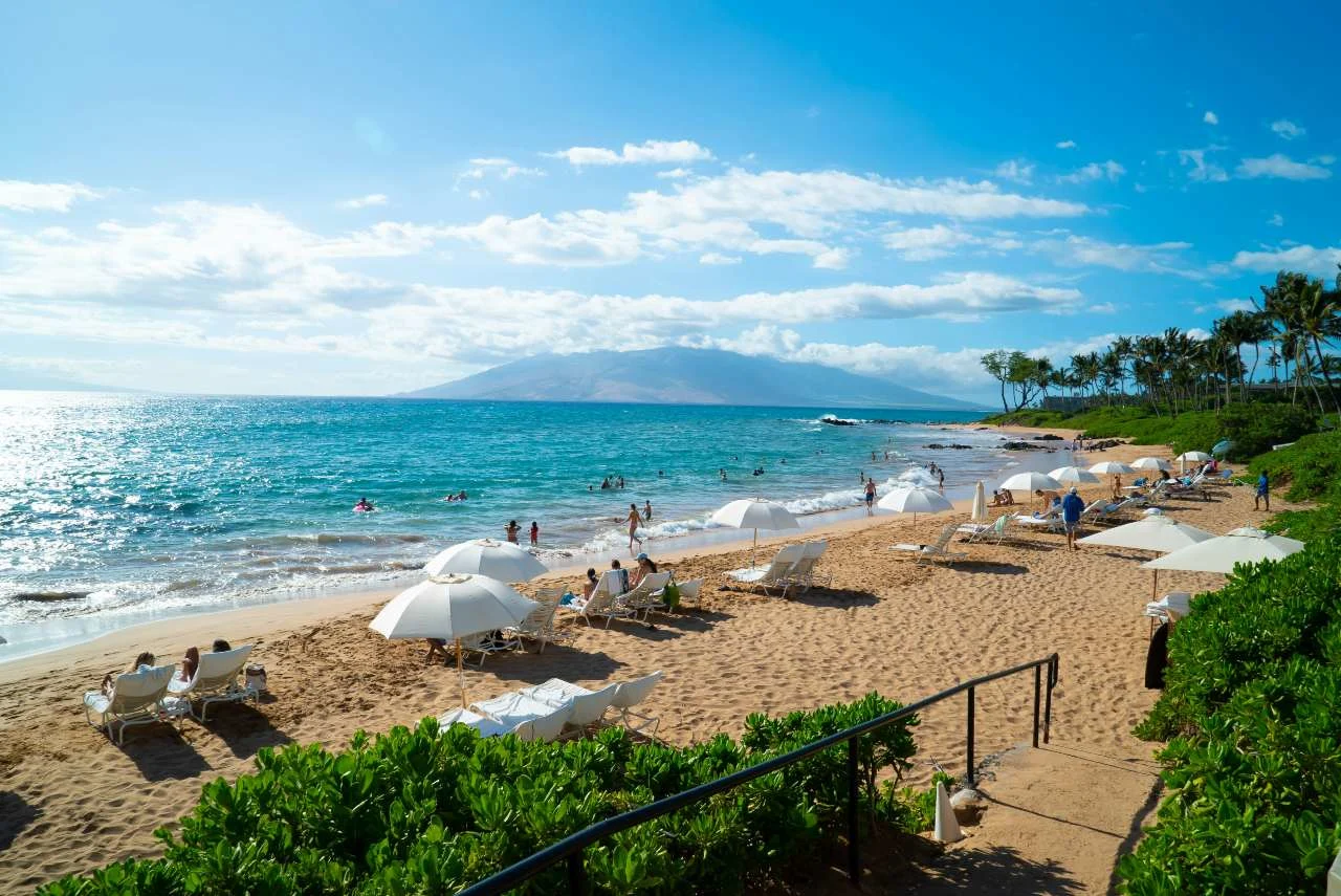 A sandy Maui beach lined with white lounge chairs and umbrellas, with swimmers in the turquoise water and a mountain silhouette across the bay