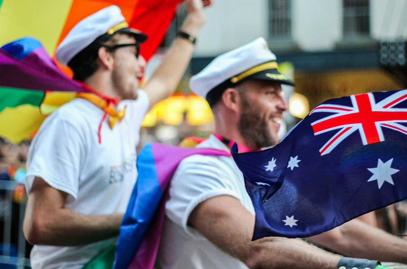 Two friends at Sydney Pride wear sailor caps and rainbow flag capes while unfurling an Australian flag. Friends in sailor caps and rainbow flag capes hold an Australian flag at Sydney Pride, which runs across two weeks in June each year.