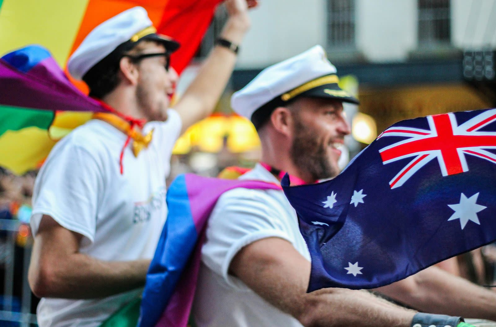 Two friends at Sydney Pride wear sailor caps and rainbow flag capes while unfurling an Australian flag. Friends in sailor caps and rainbow flag capes hold an Australian flag at Sydney Pride, which runs across two weeks in June each year.