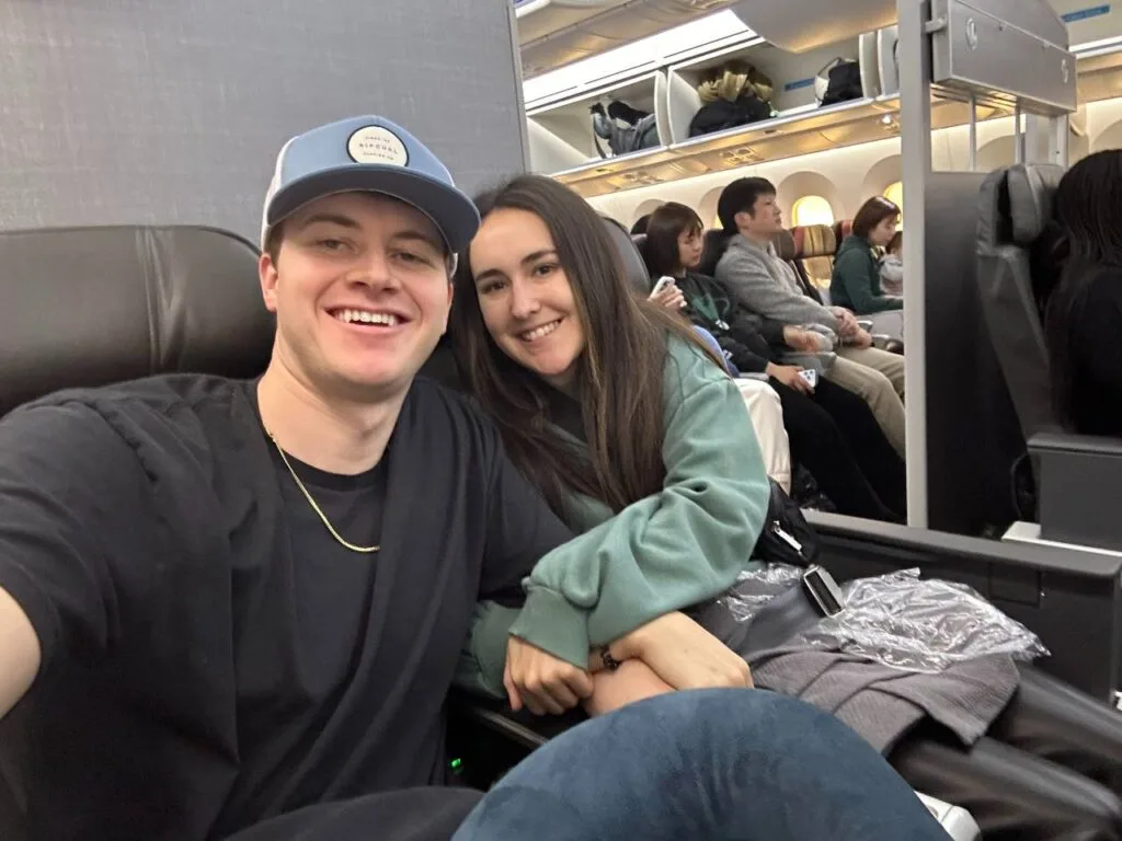 A couple smiling for a selfie in business class seats on a wide-body jet, with other passengers seated in the cabin behind them.