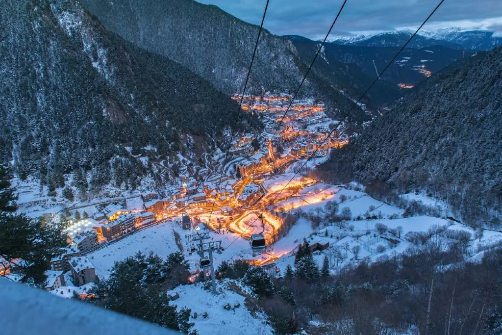 A cable car descends toward the illuminated mountain town of Andorra la Vella at dusk, surrounded by snowy peaks