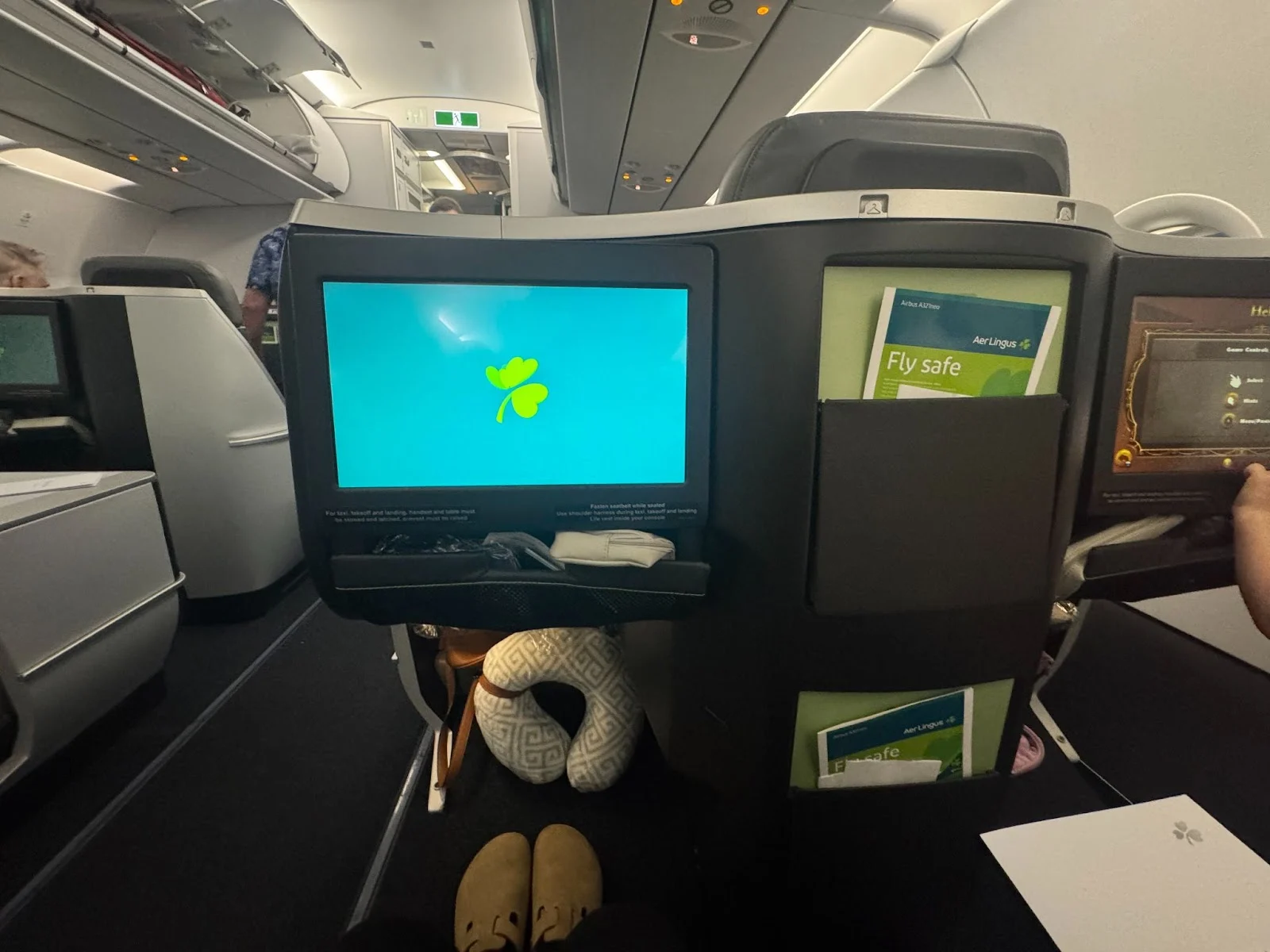 Aer Lingus business class seatback screen displaying the airline's shamrock logo, with a passenger's feet in slippers and a neck pillow below.
