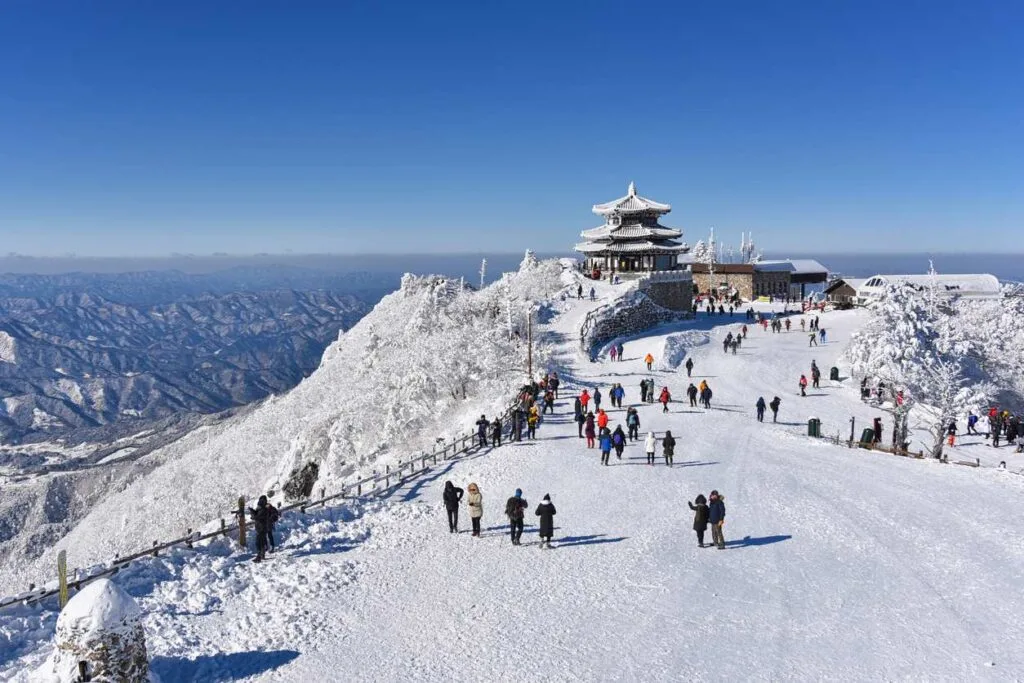 Tourists walking along a snow-covered mountain ridge toward a frosted pavilion at a South Korean ski resort summit