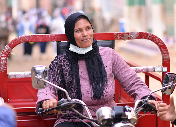 An Egyptian woman in a pink shirt and black head scarf. She smiles as she rides a motorcycle through a village.