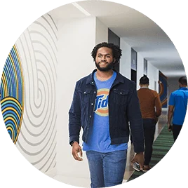 A young man in a t-shirt, jacket, and jeans walks through the hallways of his office.