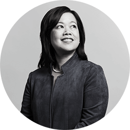 An Asian woman with shoulder-length black hair wears a dark suede jacket and a string of pearls. She smiles as she looks off in the distance. The image is black and white.