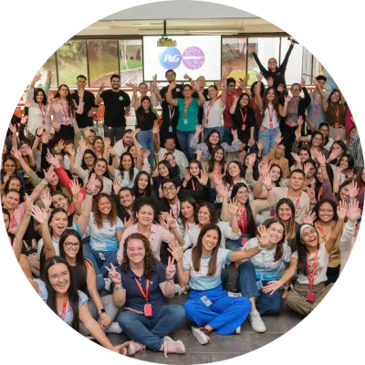 A large group of Hispanic Procter & Gamble employees pose, with many sitting on the floor and others standing in the background. They are waving and smiling. The group is predominantly female.