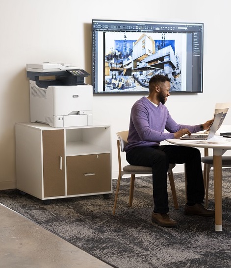 Man working at an office table with a large monitor and a Xerox VersaLink C625 MFP behind him