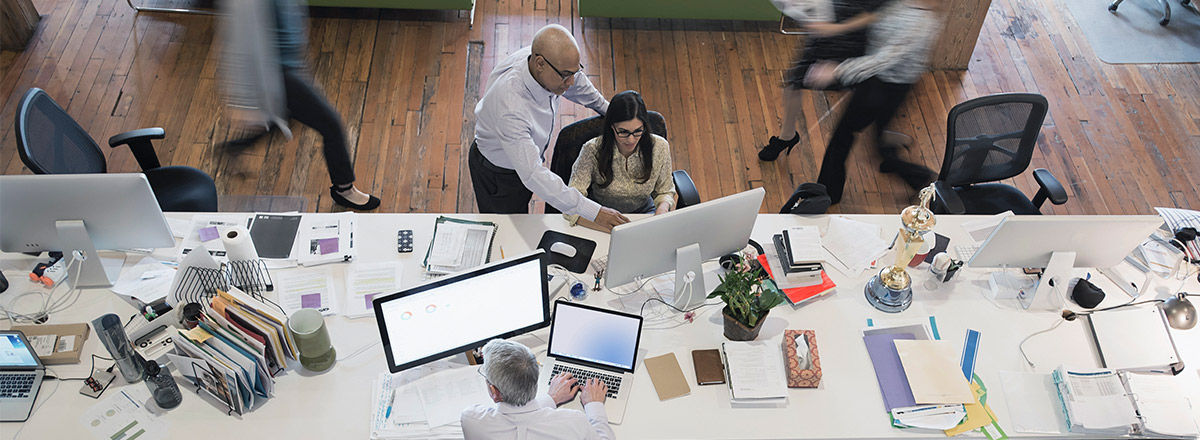 Coworkers in front of a computer, at a large desk, with people walking by