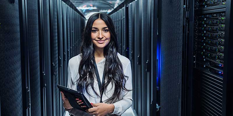 Smiling IT tech with tablet standing between rows of servers