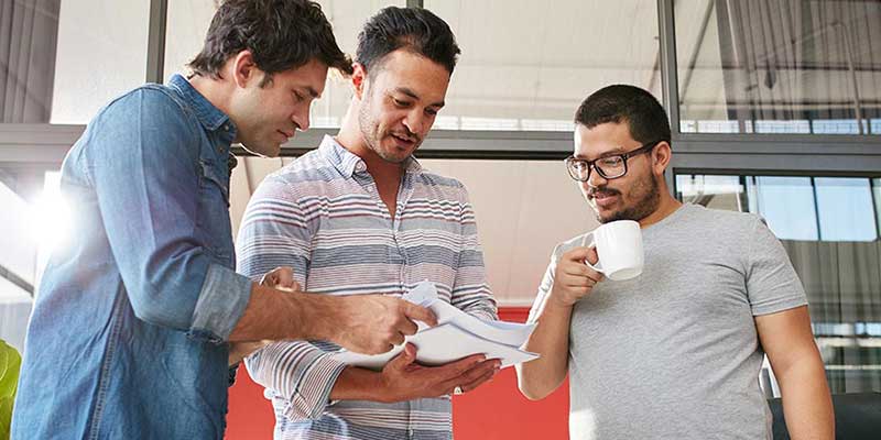 Three co-workers standing together in a hallway looking at a stack of printed documents. One is holding a cup of coffee.