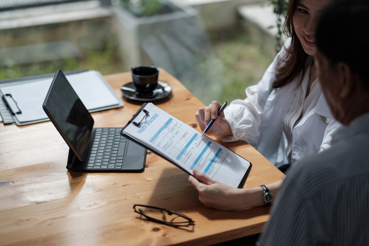 Man and woman filling out health insurance form on clipboard. The table in the background has a tablet, coffee cup, glasses, and another clipboard.