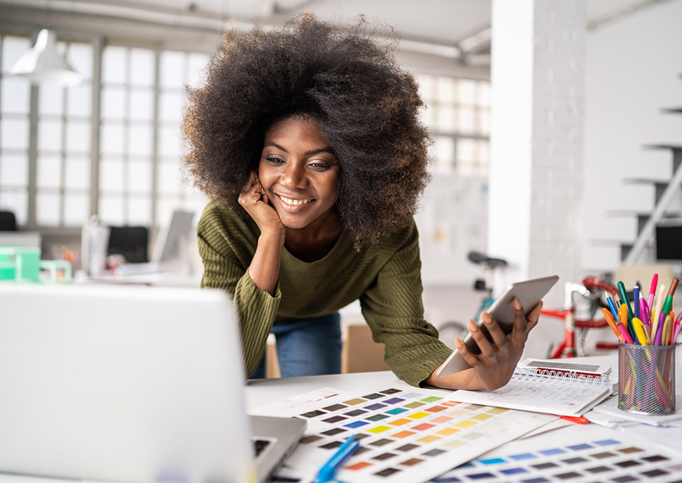 A happy woman with a laptop and tablet, looking at printed color swatches