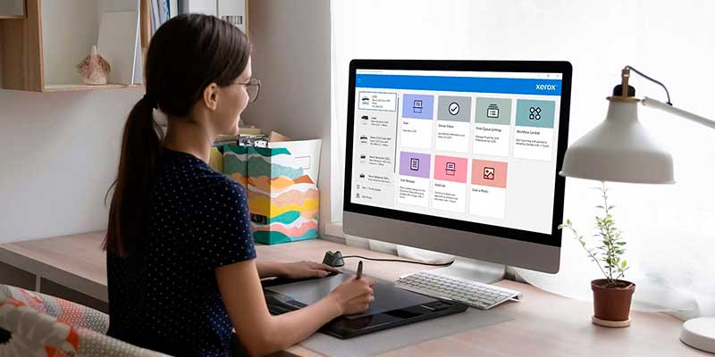 Young professional sitting at a desk by a window, working on a monitor with Xerox Print and Scan Experience App