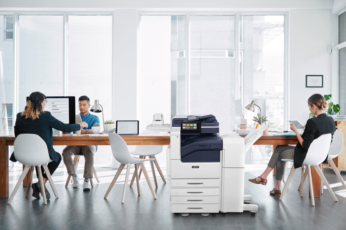 Brightly lit office with big windows, with a Xerox VersaLink C7100 MFP between people working at a table.