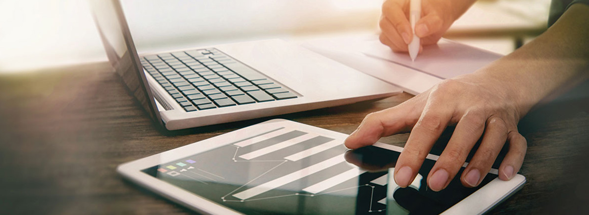 Person writing on a pad of paper while using a tablet, with a laptop nearby