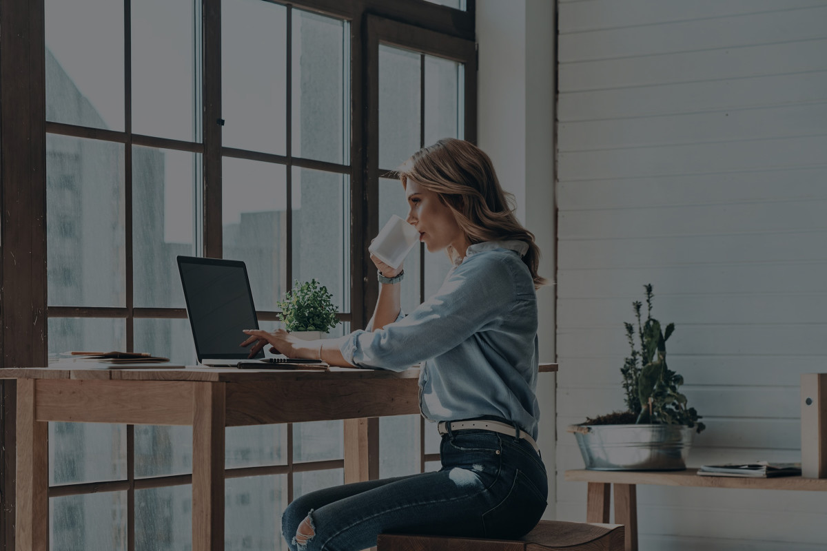 Woman working on a laptop at a desk at a window, drinking coffee