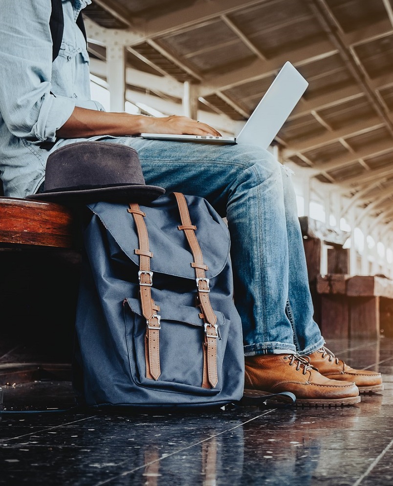 Neck down view of a person sitting in a train station with a laptop on their lap, with a backpack and fedora beside them