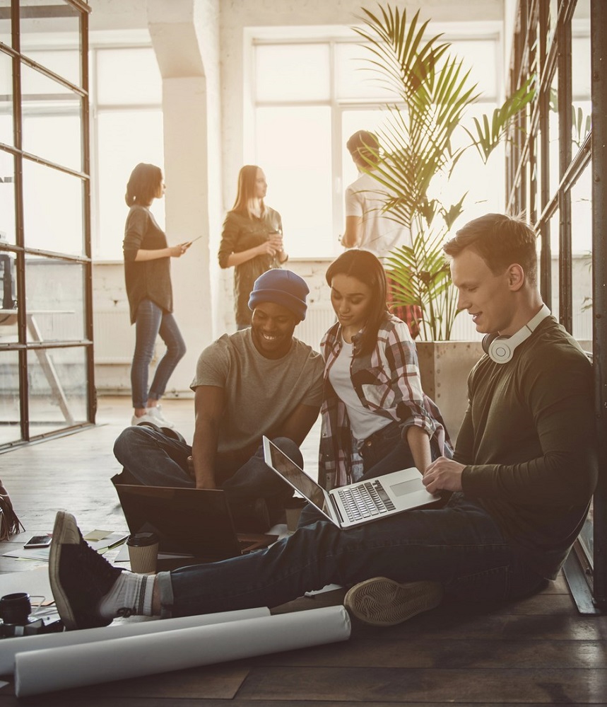 3 employees sitting on the floor looking at laptops, with 3 more employees standing behind them