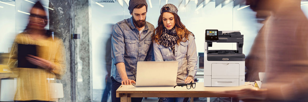 Two people with trendy hats using a laptop in front of a Xerox MFP