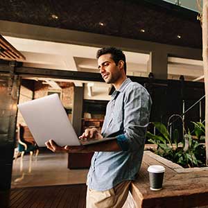 Man working in a hallway on a laptop with a cup of coffee. Plants and a stairway behind him.