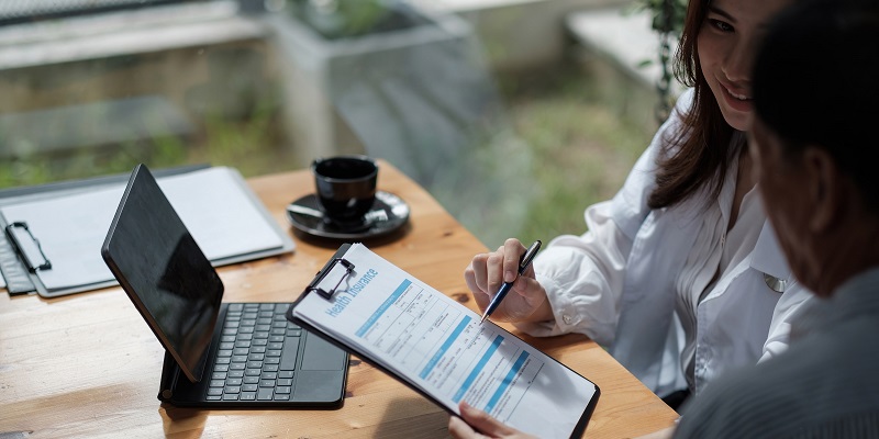 Man and woman filling out health insurance form on clipboard. The table in the background has a tablet, coffee cup, and another clipboard.