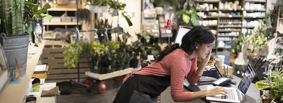 Florist wearing an apron, surrounded by lots of green plants, doing paperwork on a laptop