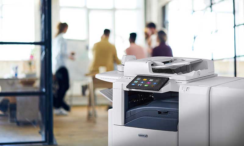 Xerox AltaLink MFP in a brightly lit office with a meeting in the background
