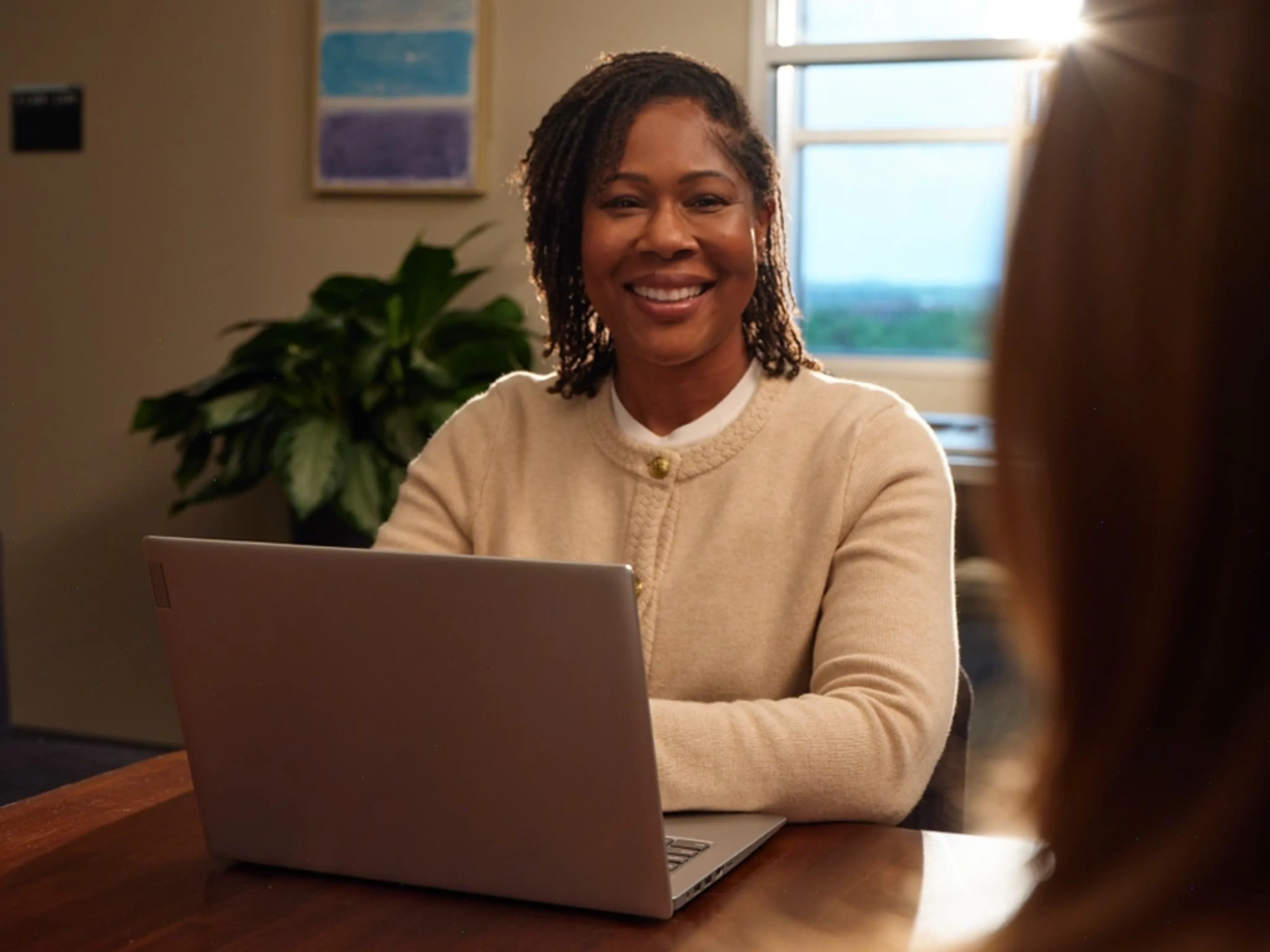 A smiling woman sitting at a desk with a laptop, engaging in a conversation.