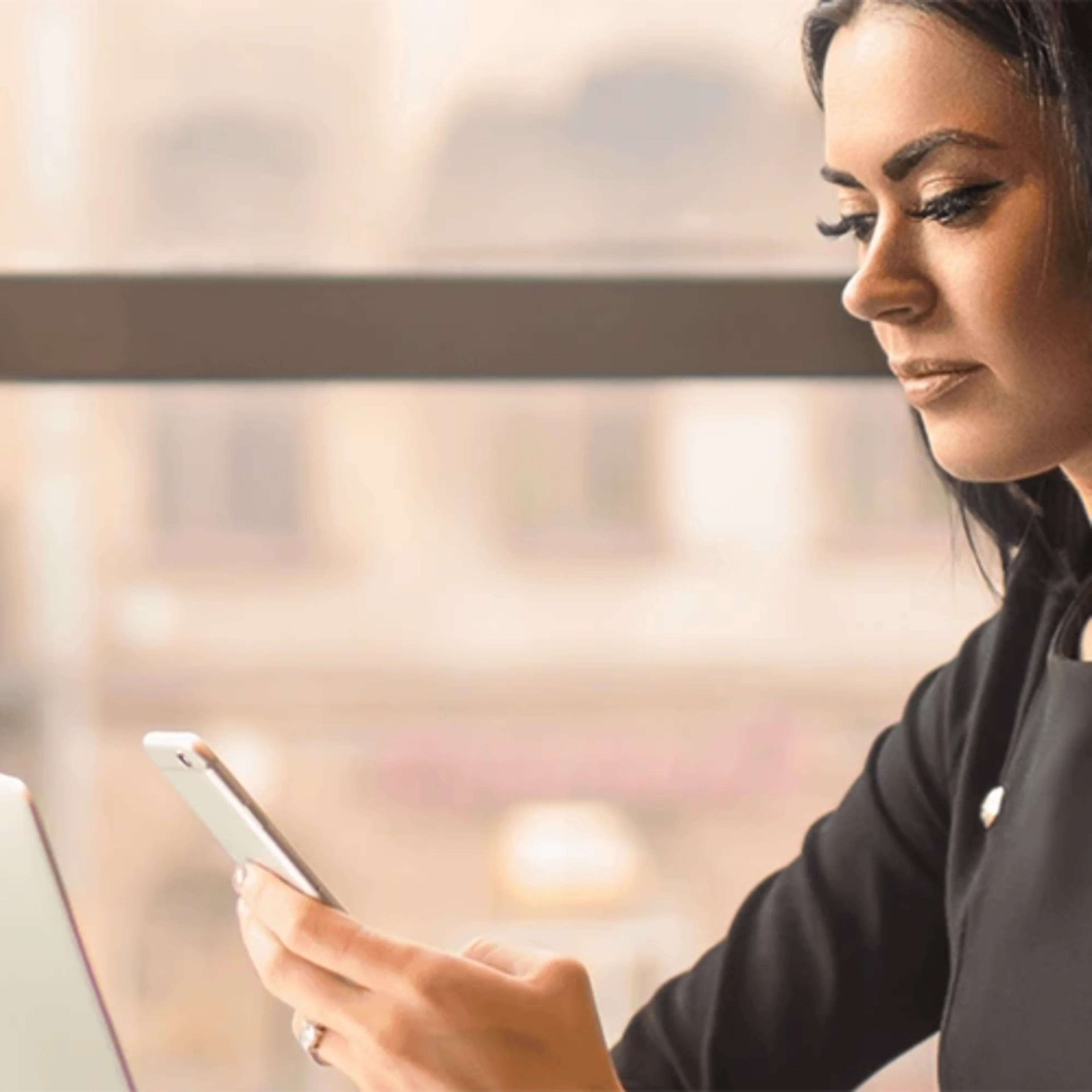 A woman looks down her mobile phone while working on her laptop.