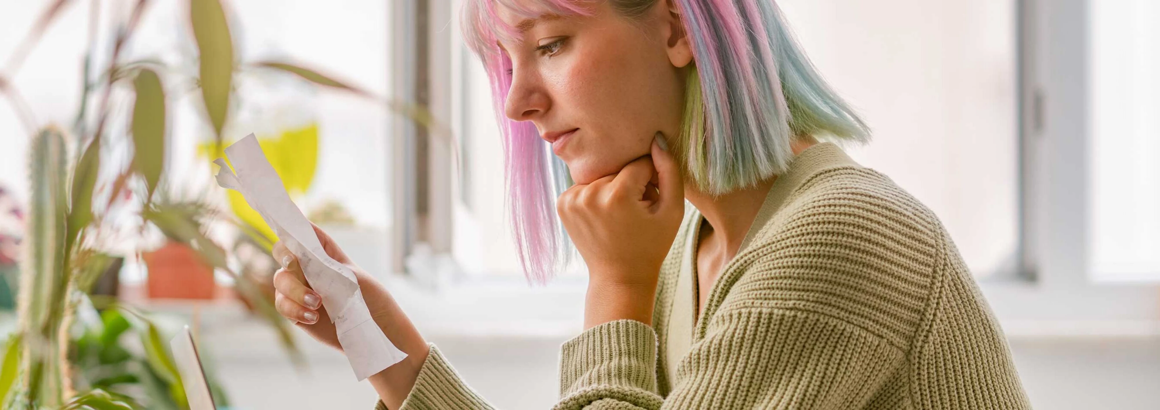 Sad looking girl with pink and colorful hair, sitting looking at a receipt with her hand on chin.