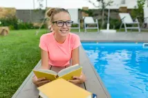 Girl lying next to a pool while holding a book and looking at the distance