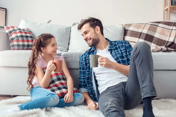 Daughter and father sitting on the floor drinking tea and looking at each other while smiling