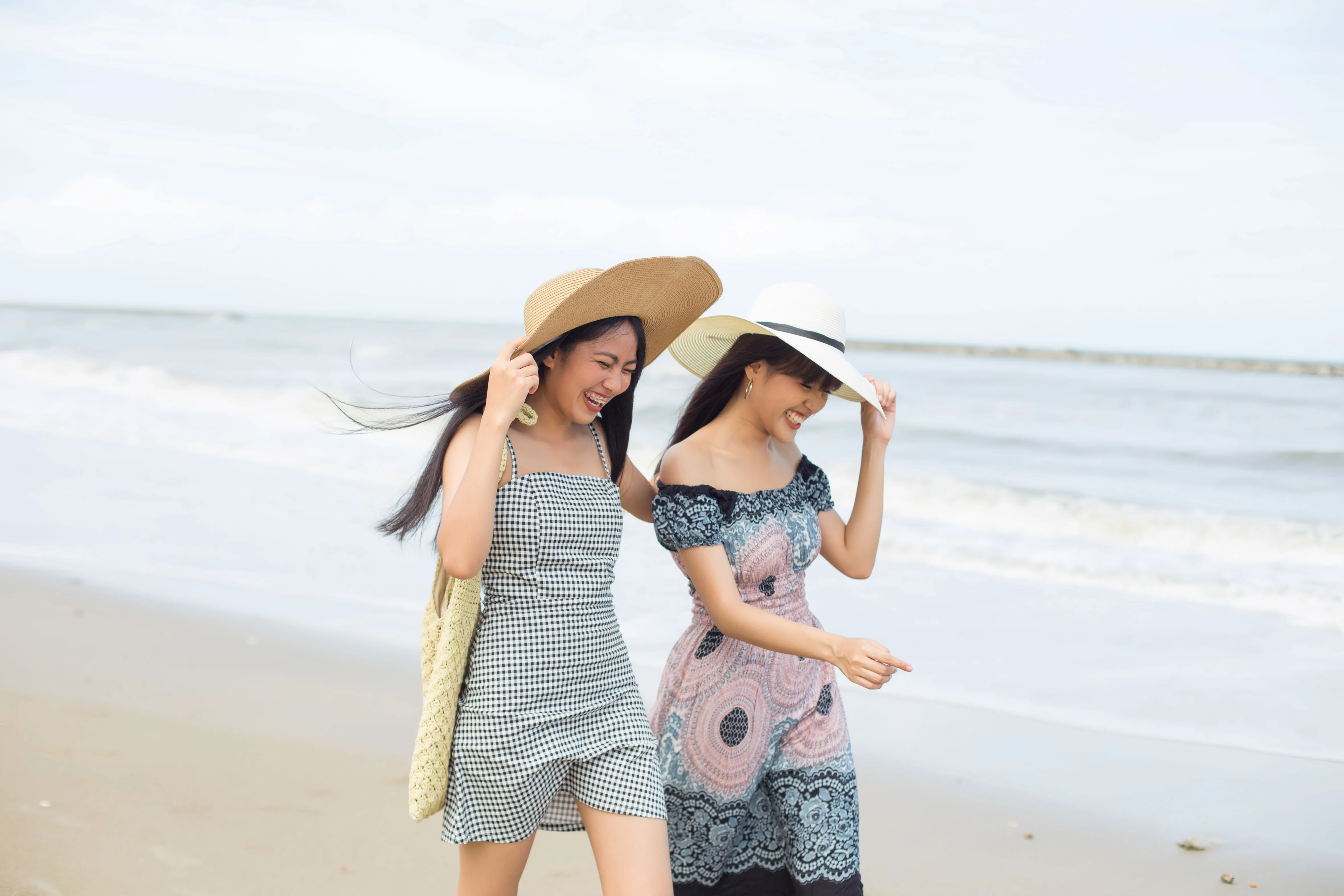 Two women laughing while holding their hats and walking on the beach