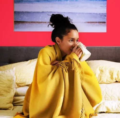 Young woman sitting on her bed with a blanket on her shoulders and sipping a cup of tea