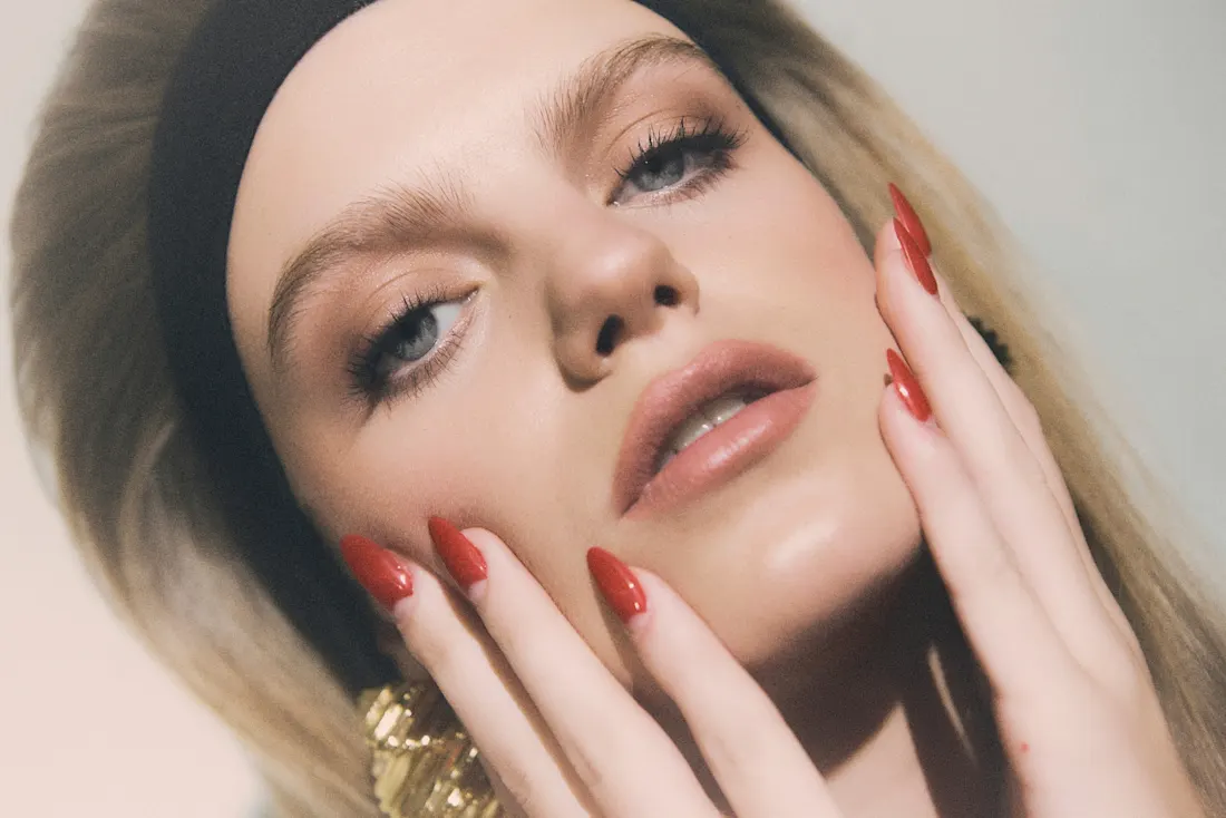 Primer plano de rostro con piel luminosa y uñas rojas apoyadas en la mejilla