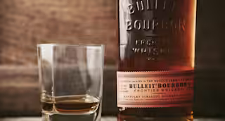 A bottle of Bulleit Bourbon sitting next to a rocks glass, in front of a mountain range with snow on it