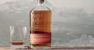 A bottle of Bulleit Bourbon sitting next to a rocks glass, in front of a mountain range with snow on it.