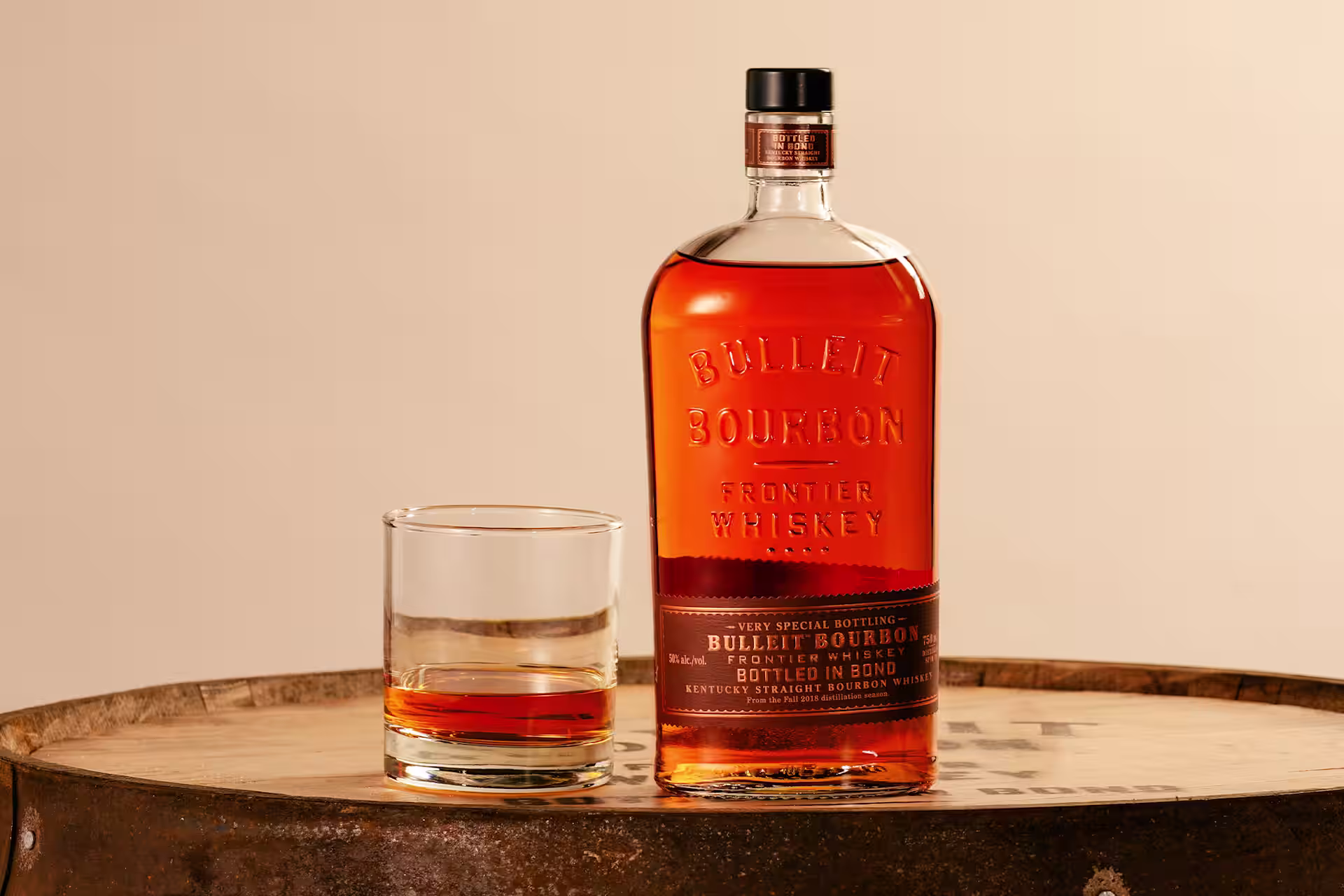 A bottle of Bulleit Bottled in Bond sitting next to a glass in front of two barrels