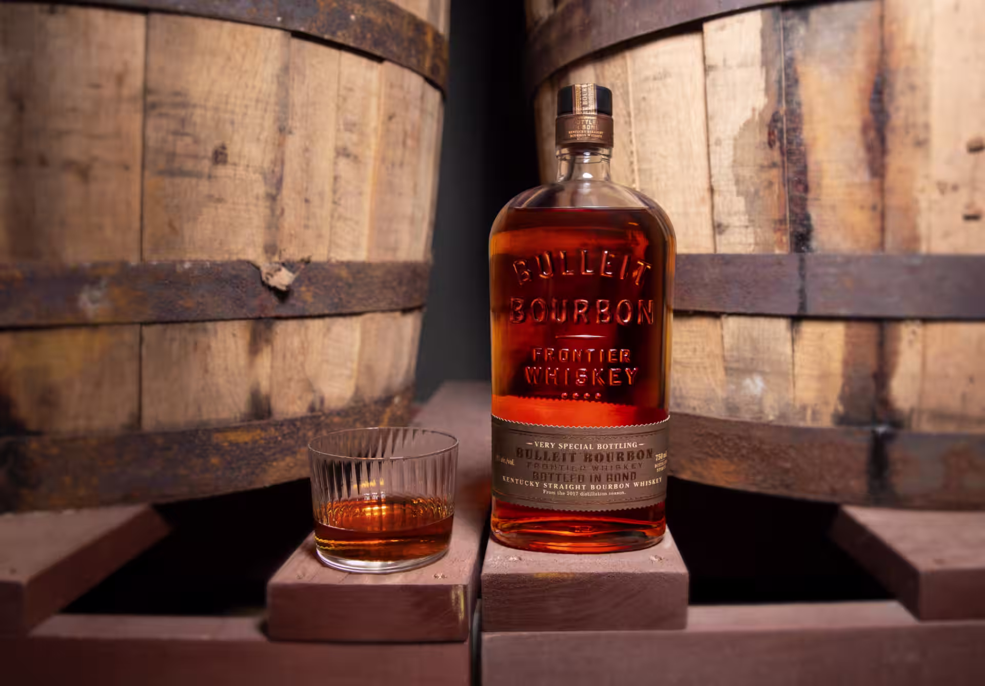 A bottle of Bulleit Bottled in Bond sitting next to a glass in front of two barrels