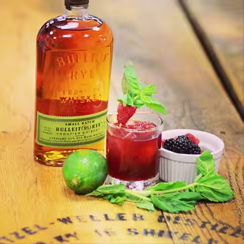 A bottle of Bulleit Rye Whiskey next to a cocktail and fruit ingredients