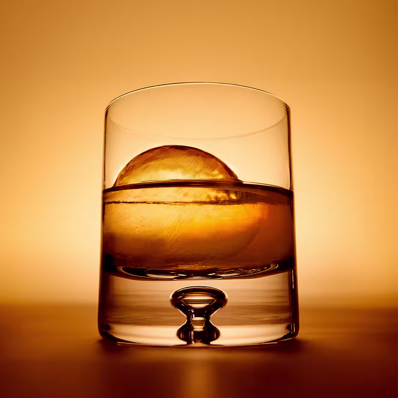 Glass of whiskey with a large sphere of ice against a warm amber background