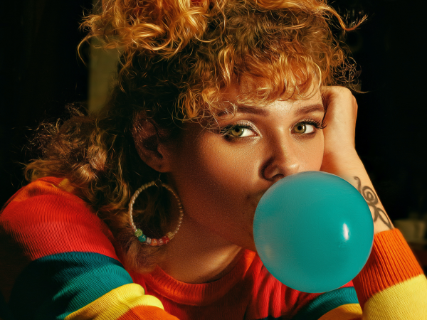Young adult woman looking at the camera while blowing a bubble with her bubble gum. The bubble is blue